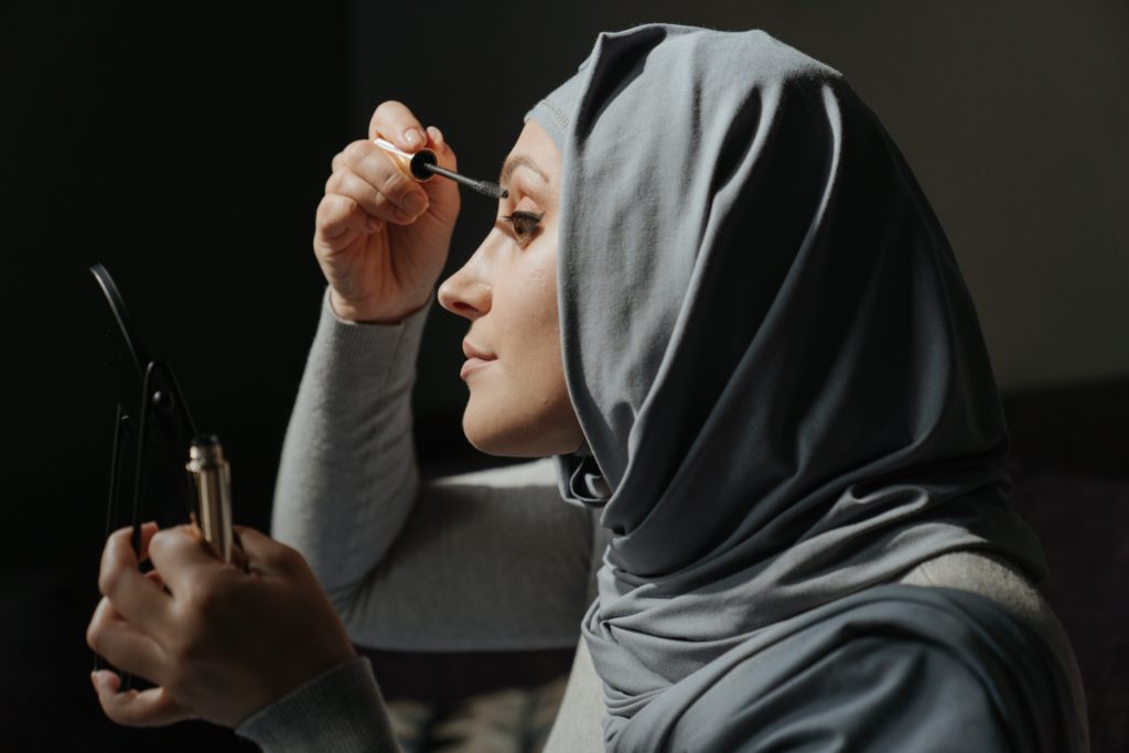 Woman in a hijab holds mirror and applies mascara