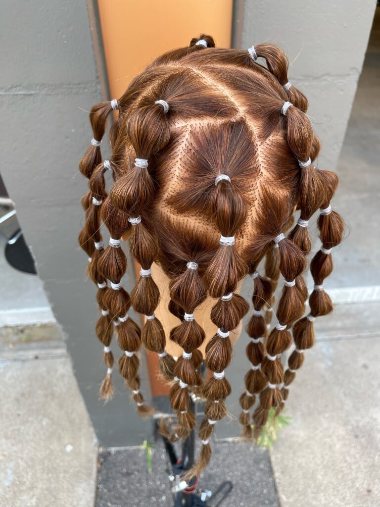 Mannequin with hair parted into geometrical sections to create pony tails that are held in place with hair elastics and banded down the length to create small bubbles. Mannequin is facing away.