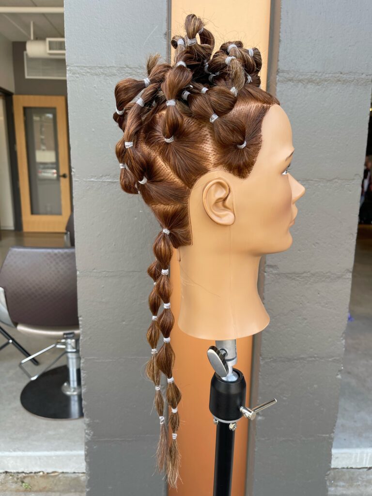 Mannequin with hair parted into geometrical sections to create pony tails that are held in place with hair elastics and banded down the length to create small bubbles.  The top half of these ponytails are piled on top of the head. Mannequin is facing right.