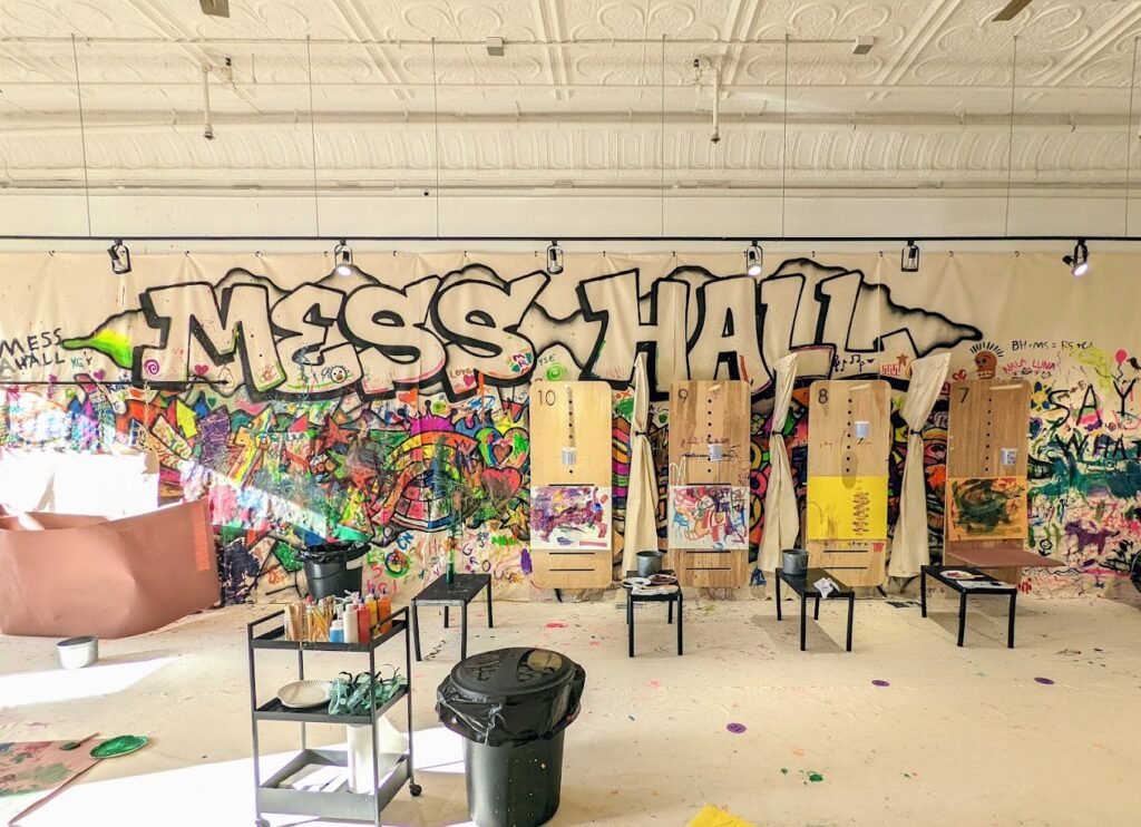 A sunny view of Mess Hall's painting room featuring plywood easels, abstract paintings, paints and supplies, and a backsplash with the words "Mess Hall" written in graffiti script and covered in paint.