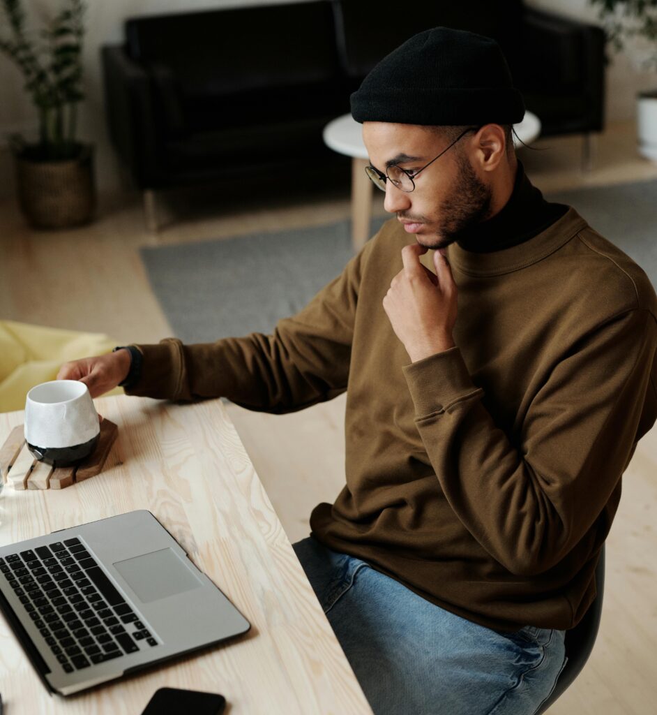 Man in black beanie and brown sweater works at a laptop and holds a small cup of coffee