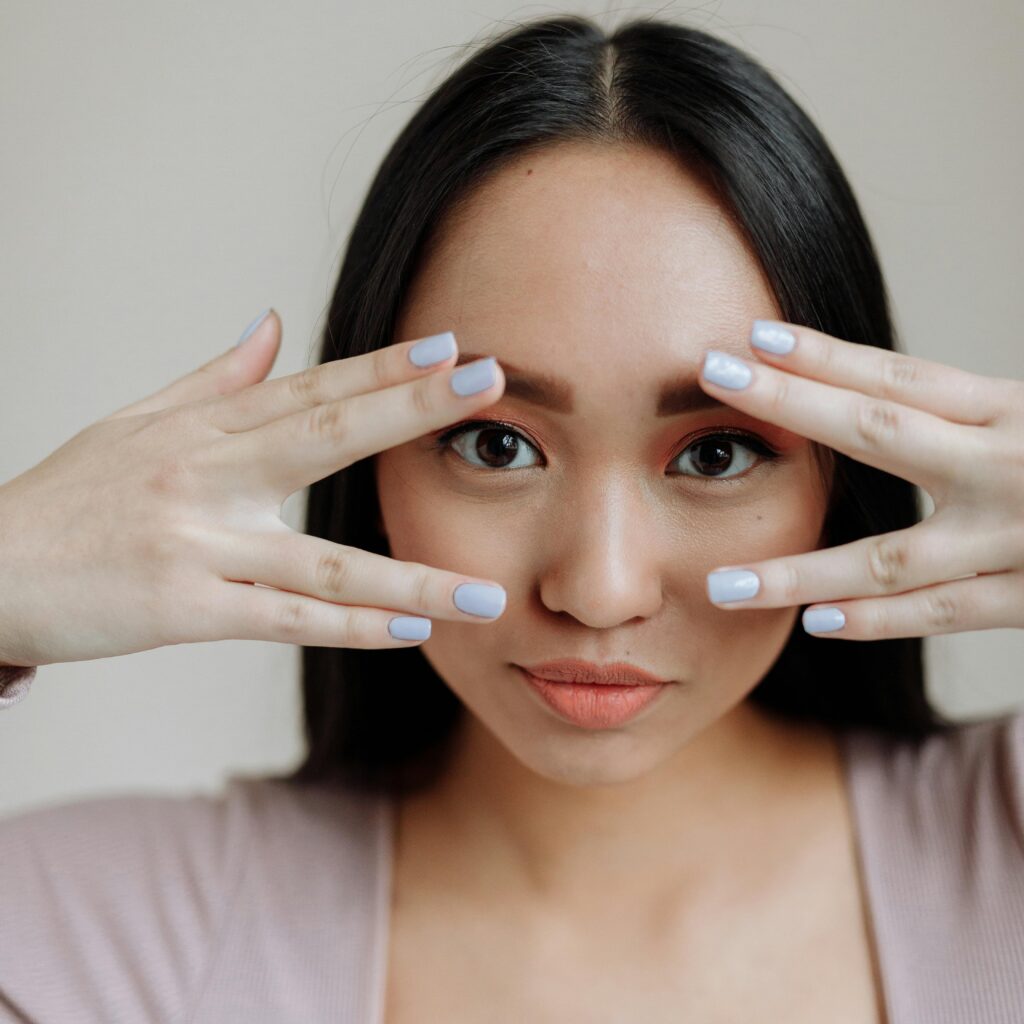 Closeup of young woman's face.  She is holding both hands in front of her eyes so you can see her nails, but she is looking through her fingers so you can see her eyes as well. Her fingernails have grown past the tips of her fingers and are painted pastel blue. 