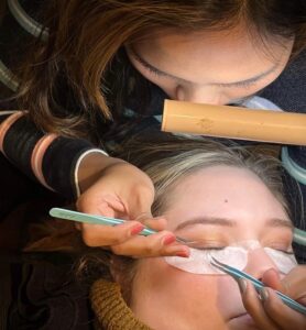 Student applies lash extensions to a client, illuminated by a lamp