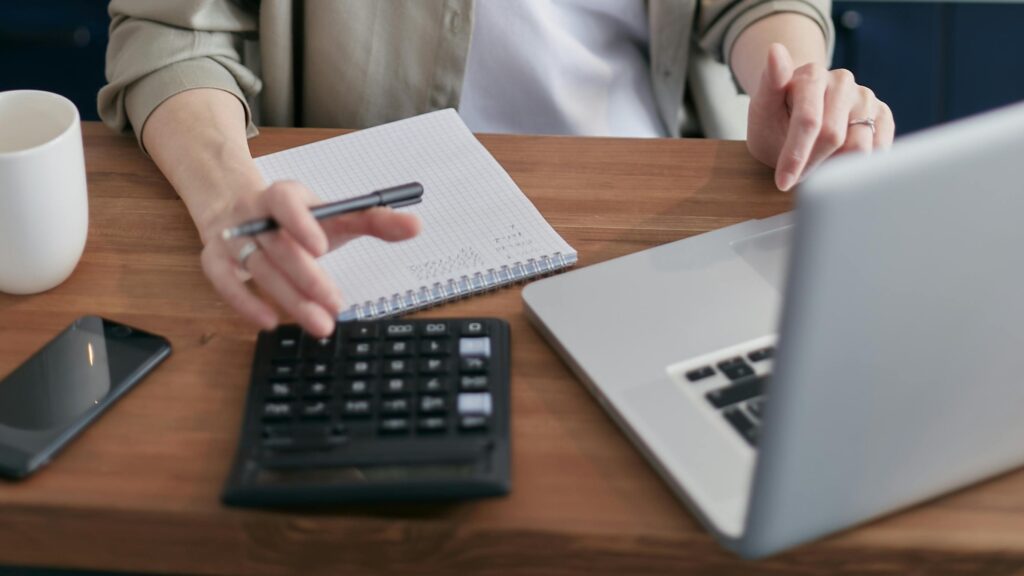 Someone sits at a laptop with a paper and calculator to calculate out expenses.