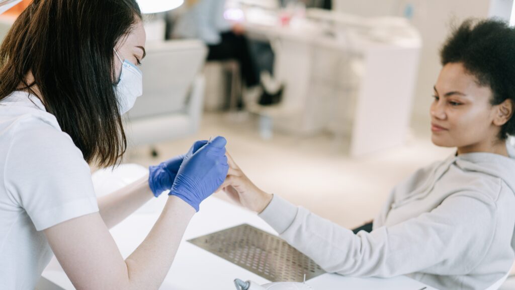 A smiling nail technician wearing a mask works on a client's nails.