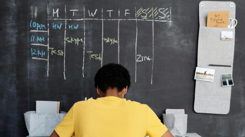A student sits at a desk with a schedule written behind them on a blackboard.
